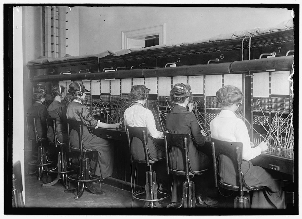 Six operators, all women, all wearing headets, sitting in front of a telephone exchange. 