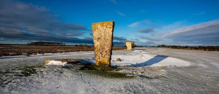 Kungsstenarna på Öland. Två stående stenar, en i förgrunden och en längre bort. Snö på marken och en blå himmel med några måln långt borta. Solen skiner på stenarna och skuggan från närmsta stenen faller snett bortåt åt höger.