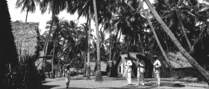 Tre kostymklädda män bland palmer och hus. Till vänster syns ett barn. Cabedello, Brasilien.